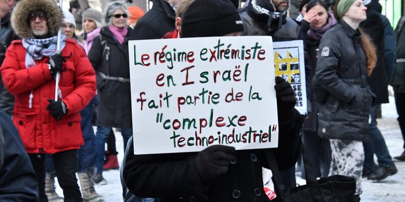 Une manifestation à Montréal pour Palestine. Des manifestants tiennent une pancarte sur laquelle il est écrit "Le régime extrémiste en Israël fait partie de la complexe techno-industriel".